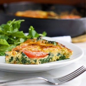 A slice of vegetable frittata with visible spinach and tomato sits on a white plate next to a small serving of fresh greens, with a fork in the foreground and a skillet in the background.