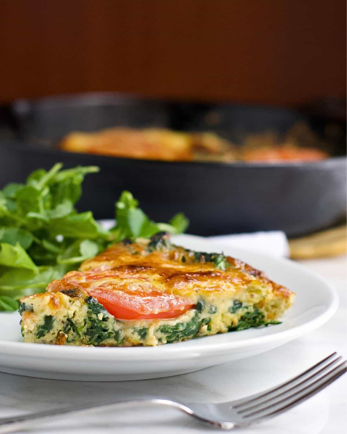 A slice of vegetable frittata with visible spinach and tomato is served on a white plate next to fresh greens, with a fork in front and a cast iron skillet in the blurred background.