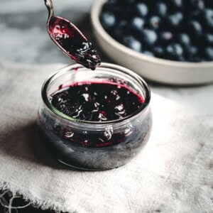 A jar of dark purple Blueberry Compote sits on a cloth with a spoonful of compote above it; a bowl of fresh blueberries is blurred in the background.