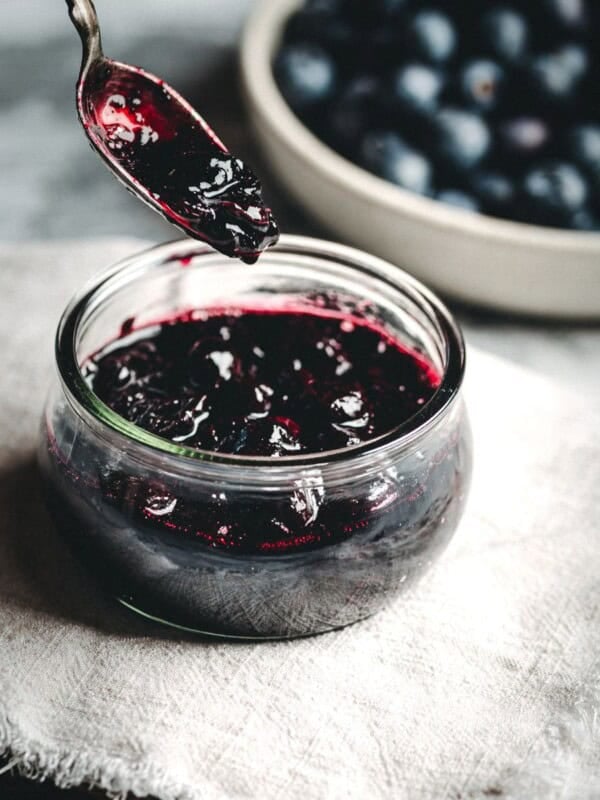 A jar of dark purple Blueberry Compote sits on a cloth with a spoonful of compote above it; a bowl of fresh blueberries is blurred in the background.