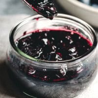 A spoon holding thick blueberry compote is held above a glass jar filled with more compote; a bowl of fresh blueberries sits blurred in the background. The text reads "Blueberry Compote, pinchandswirl.com.