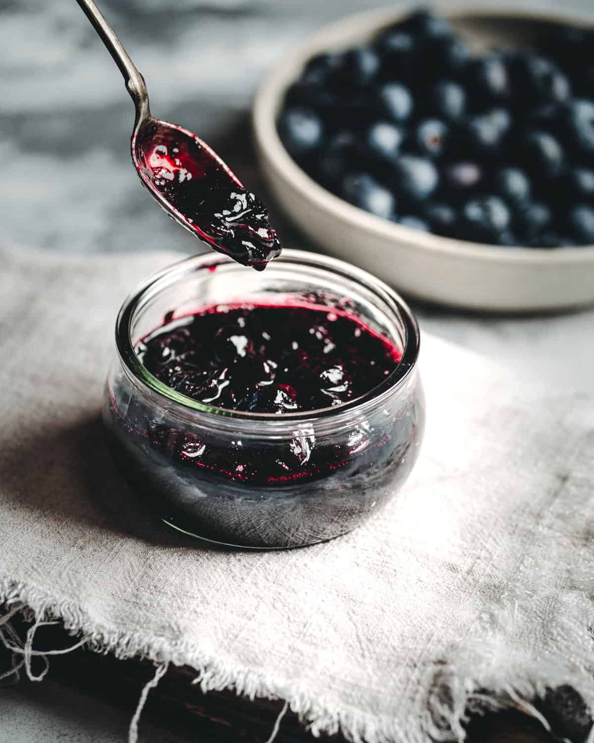 A glass jar filled with dark blueberry compote sits on a cloth, with a spoon holding jam above it. In the background, a bowl of fresh blueberries is slightly out of focus.