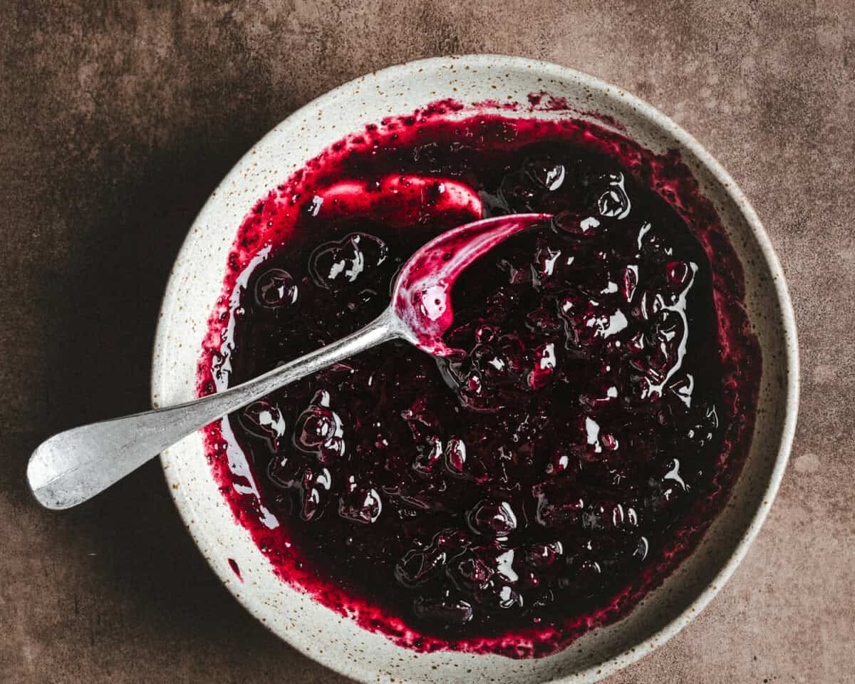 A speckled ceramic bowl filled with thick, dark blueberry compote sits on a brown surface. A metal spoon rests in the bowl, partially covered with the glossy fruit mixture.