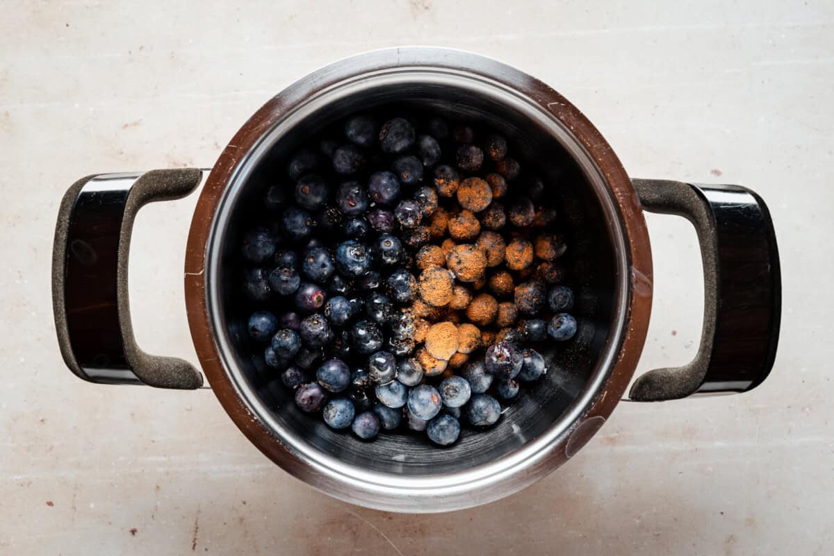 A metal pot filled with fresh blueberries and a pinch of ground cinnamon on top, viewed from above on a light-colored surface.