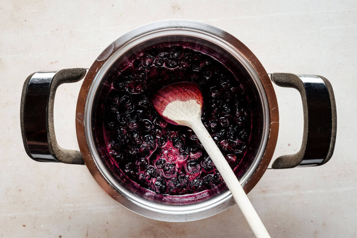 A pot filled with dark purple blueberry mixture being stirred with a wooden spoon, viewed from above on a light-colored surface.