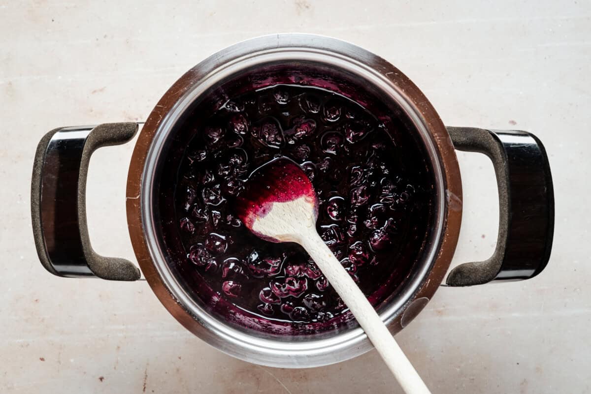 A pot of homemade blueberry compote being stirred with a wooden spoon, showing dark purple, glossy berries and syrup against a light background.