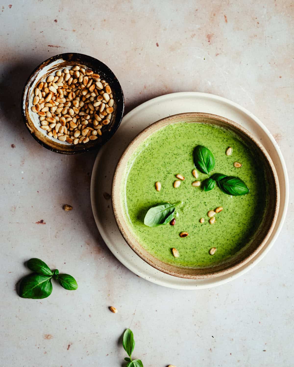 A bowl of creamy pesto sauce garnished with basil leaves and pine nuts sits on a plate, next to a small bowl filled with toasted pine nuts. Loose basil leaves and pine nuts are scattered on a light surface.