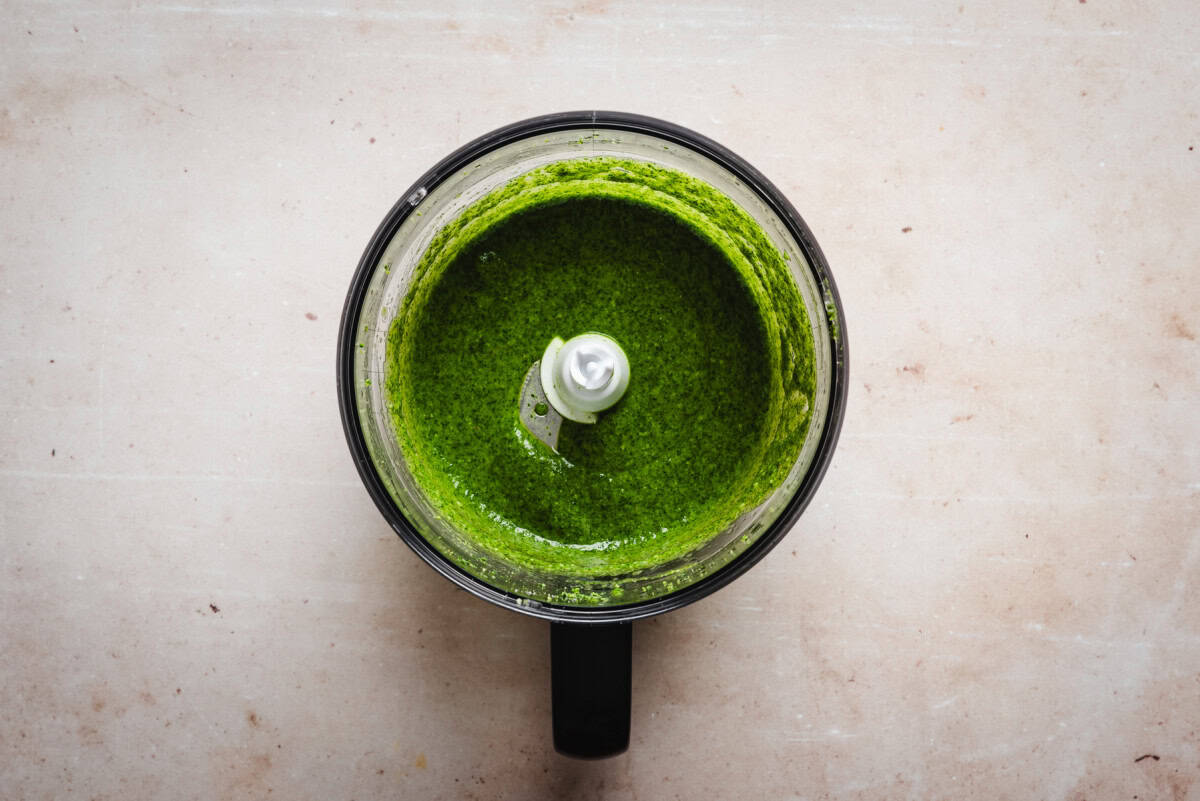 Overhead view of a food processor containing vibrant green pesto sauce, with the blade visible in the center, on a light beige surface.