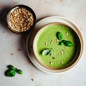 A bowl of creamy pesto sauce garnished with fresh basil leaves and pine nuts, next to a small bowl of pine nuts, on a light marble surface.
