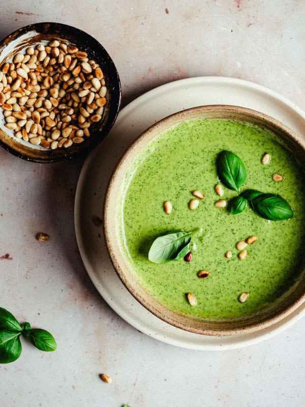 A bowl of creamy pesto sauce garnished with fresh basil leaves and pine nuts, next to a small bowl of pine nuts, on a light marble surface.
