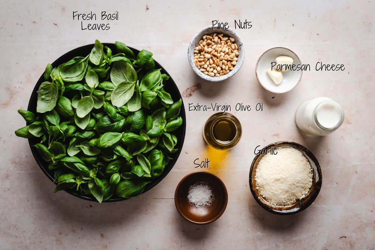 A top view of ingredients for creamy pesto sauce: a bowl of fresh basil leaves, a small bowl of pine nuts, a cup of grated parmesan cheese, a bottle of extra-virgin olive oil, a small bowl of salt, and two garlic cloves.