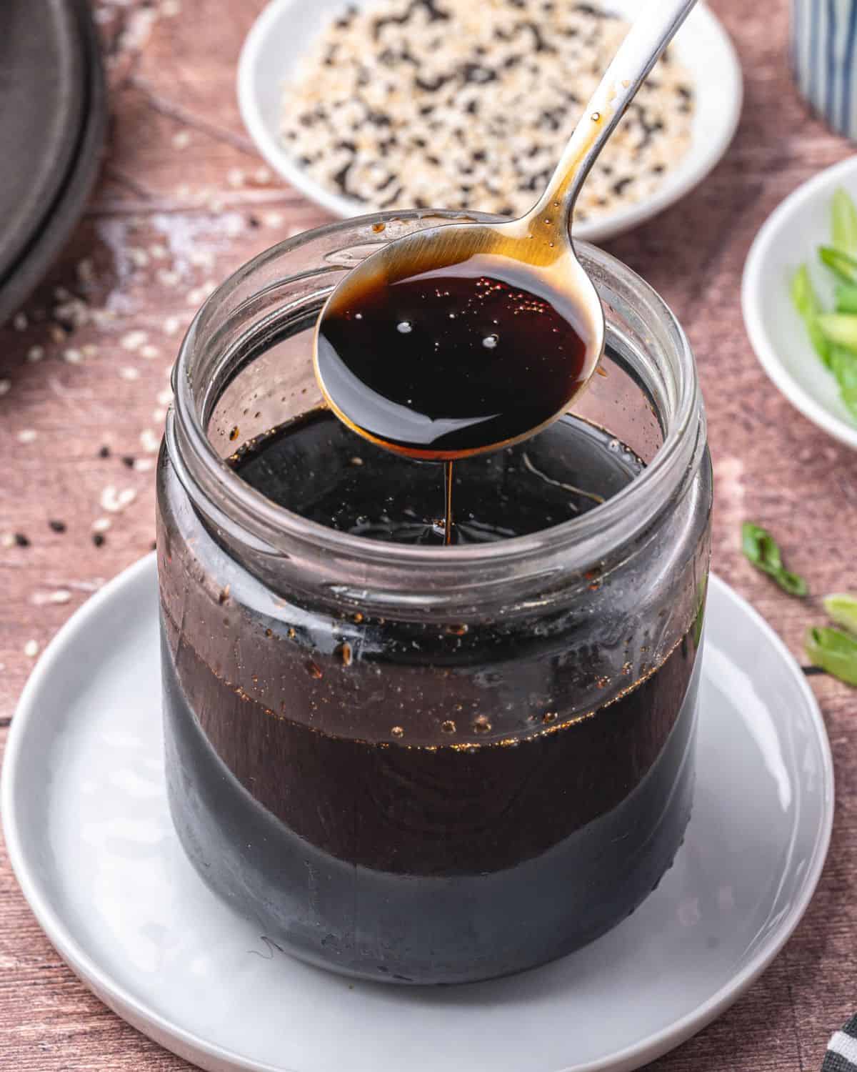 A spoon holds up a dark, glossy eel sauce above a glass jar filled with the same sauce. The jar sits on a white plate, with bowls of sesame seeds and sliced green onions in the background.