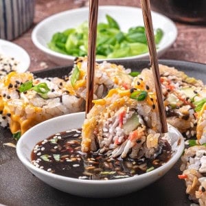 Close-up of chopsticks holding a sushi roll dipped in soy sauce, with more sushi rolls on a plate, garnished with sesame seeds and green onions. Sliced cucumbers and dipping sauce are in the background.