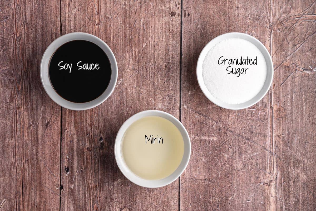 Three small bowls on a wooden surface containing soy sauce, granulated sugar, and mirin, each labeled with text inside the bowls.