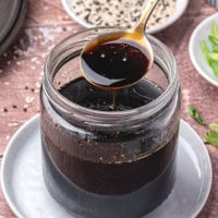 A jar of dark soy sauce sits on a white plate, with a spoon lifting some sauce above the jar. In the background are sesame seeds and sliced green onions on a wooden surface.