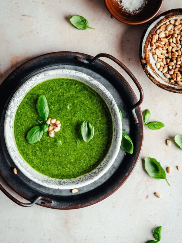 A bowl of fresh green basil pesto sauce garnished with basil leaves and pine nuts sits on a dark tray. Nearby are scattered basil leaves, a bowl of pine nuts, and a small dish of salt on a light-colored surface.