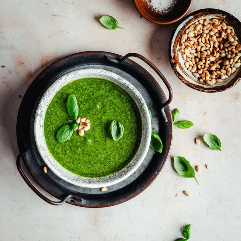 A bowl of fresh green basil pesto sauce garnished with basil leaves and pine nuts sits on a dark tray. Nearby are scattered basil leaves, a bowl of pine nuts, and a small dish of salt on a light-colored surface.