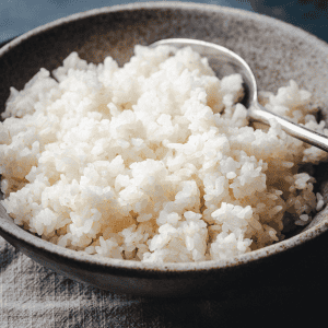 A rustic brown bowl filled with steaming sushi rice, with a metal spoon resting on the side of the bowl. The bowl sits on a textured cloth surface.