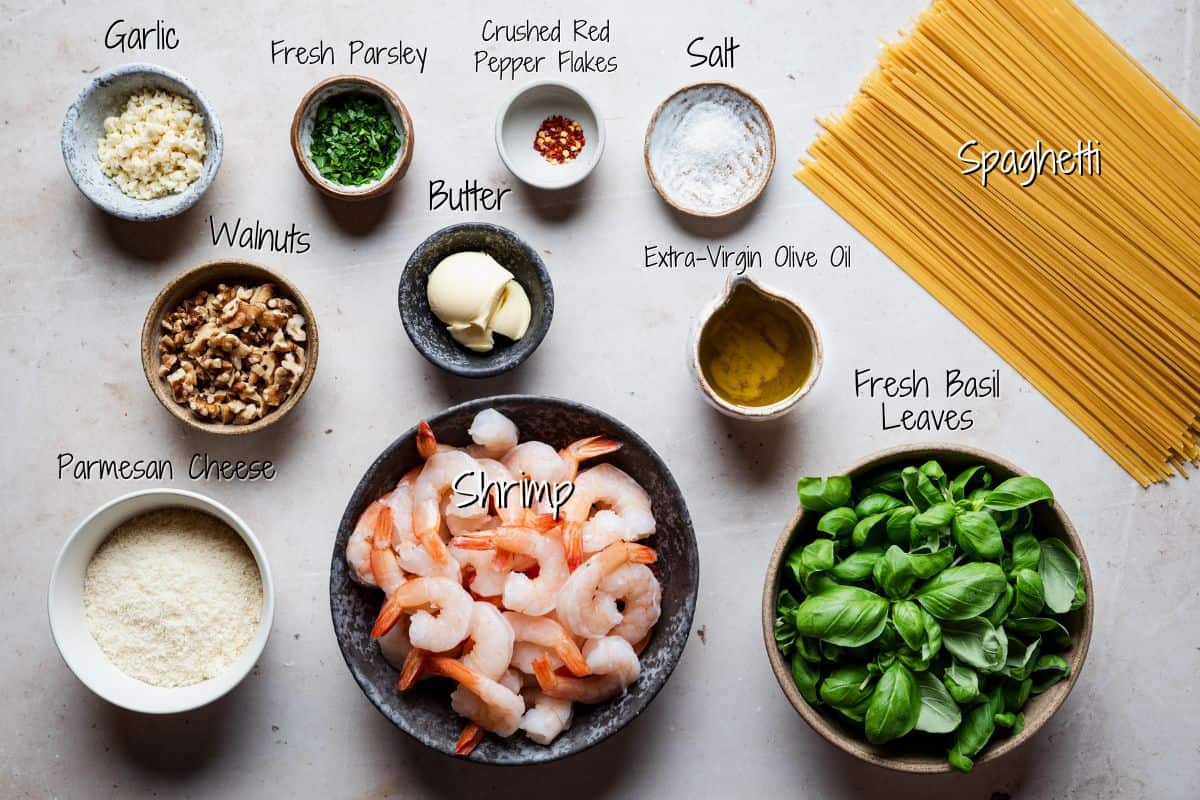 A top view of ingredients for shrimp pesto pasta, including spaghetti, shrimp, fresh basil, Parmesan cheese, walnuts, parsley, garlic, butter, red pepper flakes, salt, and olive oil, all labeled.
