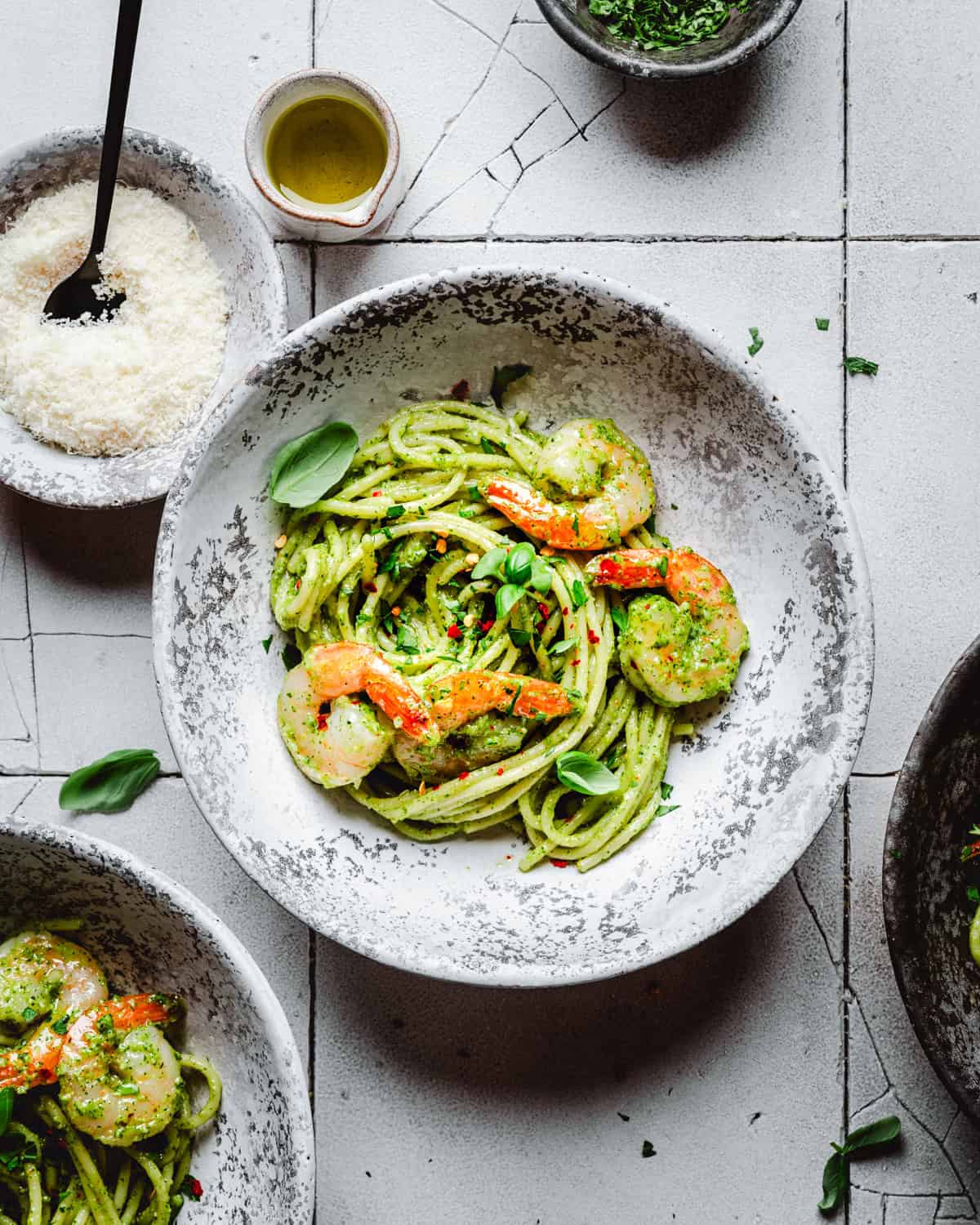A bowl of pasta tossed in green pesto sauce, topped with shrimp and garnished with fresh herbs, sits on a textured white surface, surrounded by bowls of grated cheese, sauce, and olive oil.