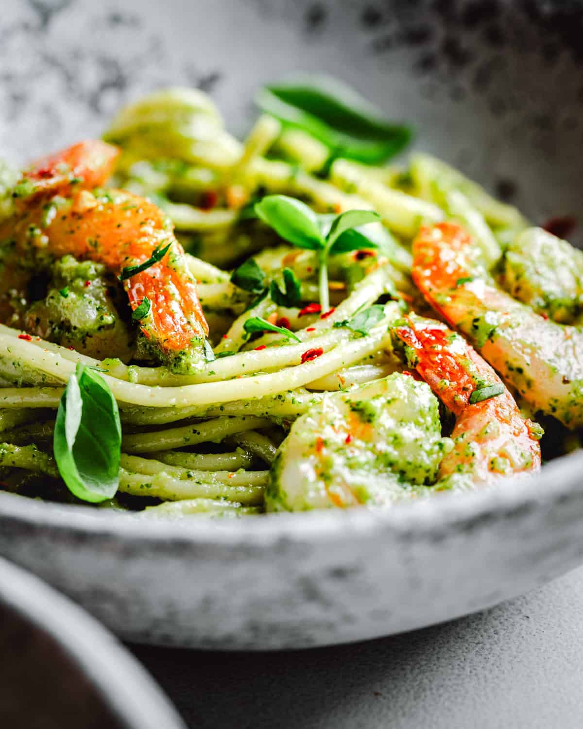 Close-up of a bowl of pasta tossed with pesto sauce, shrimp, and garnished with fresh basil leaves. The dish is vibrant with green and orange colors and served in a rustic, gray bowl.