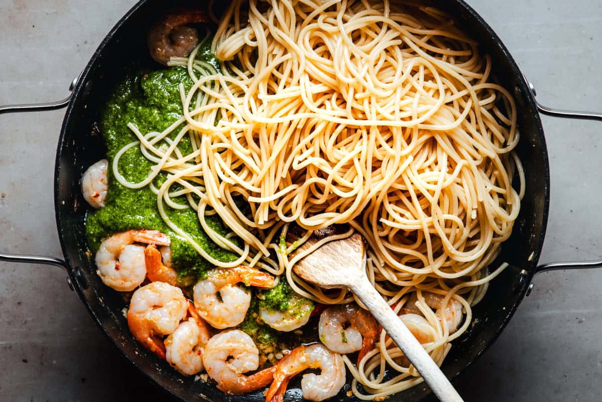 A pan filled with cooked spaghetti, sautéed shrimp, and green pesto sauce, with a wooden spoon resting in the mixture.