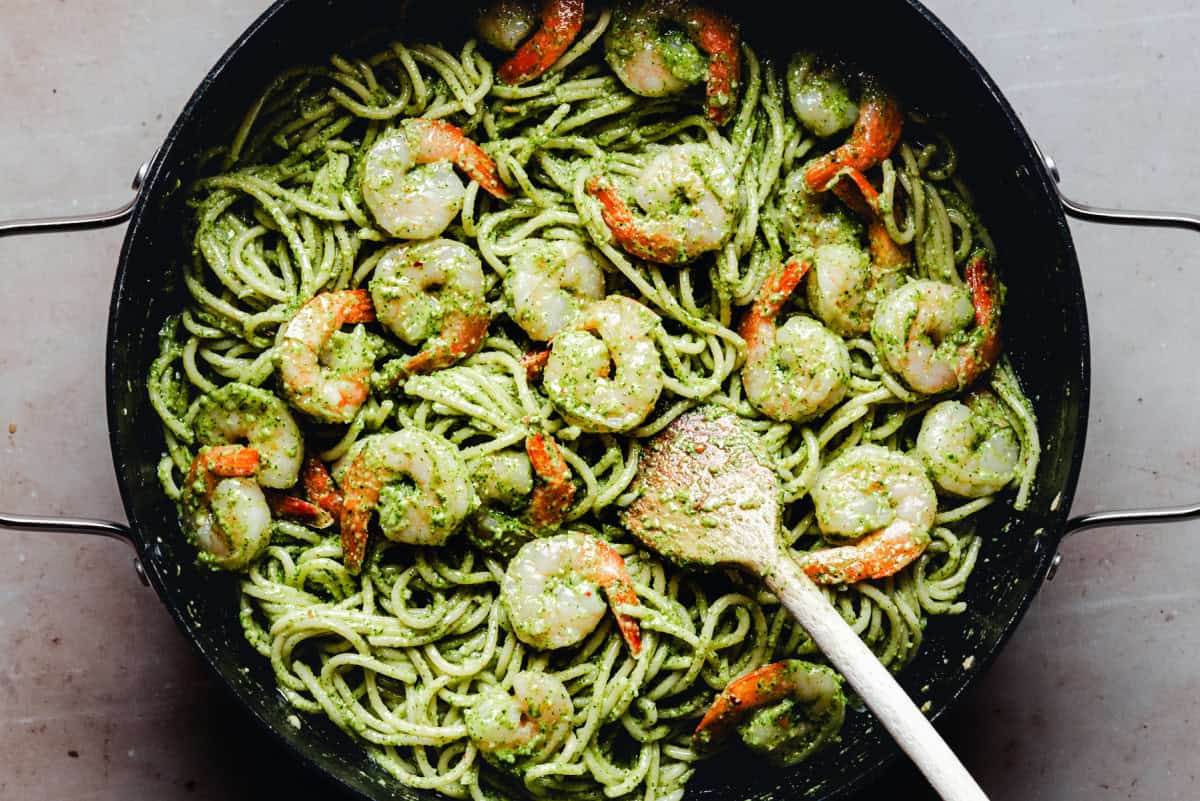 A pan filled with spaghetti coated in green pesto sauce, mixed with cooked shrimp. A wooden spoon rests in the pasta, ready for serving.