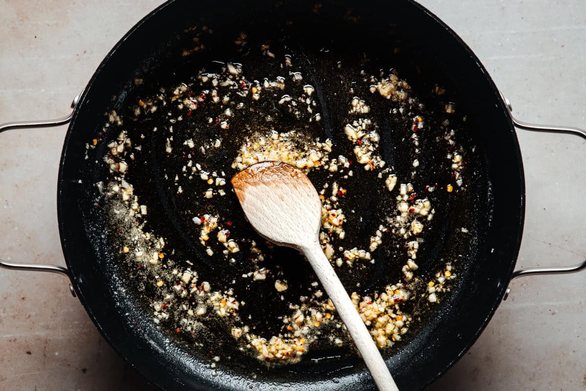 Chopped garlic and spices are being sautéed in oil in a black pan, with a wooden spoon resting inside. The pan sits on a light-colored surface.