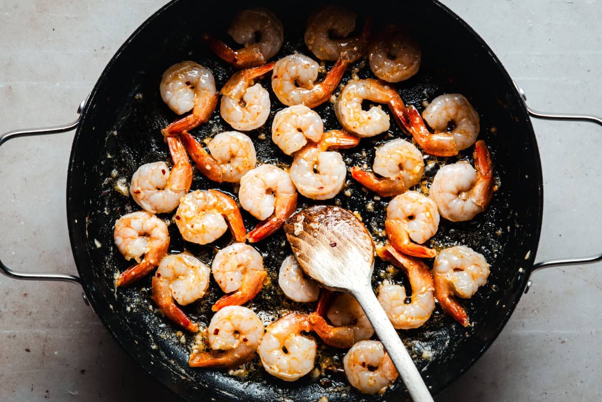 Shrimp cooking in a black skillet with seasonings, garlic, and oil, being stirred with a wooden spoon. The shrimp are pink and arranged evenly in the pan.