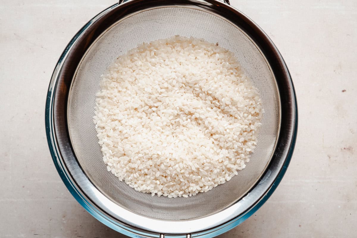 A metal strainer filled with uncooked white rice sits over a metal bowl on a light-colored surface, viewed from above.