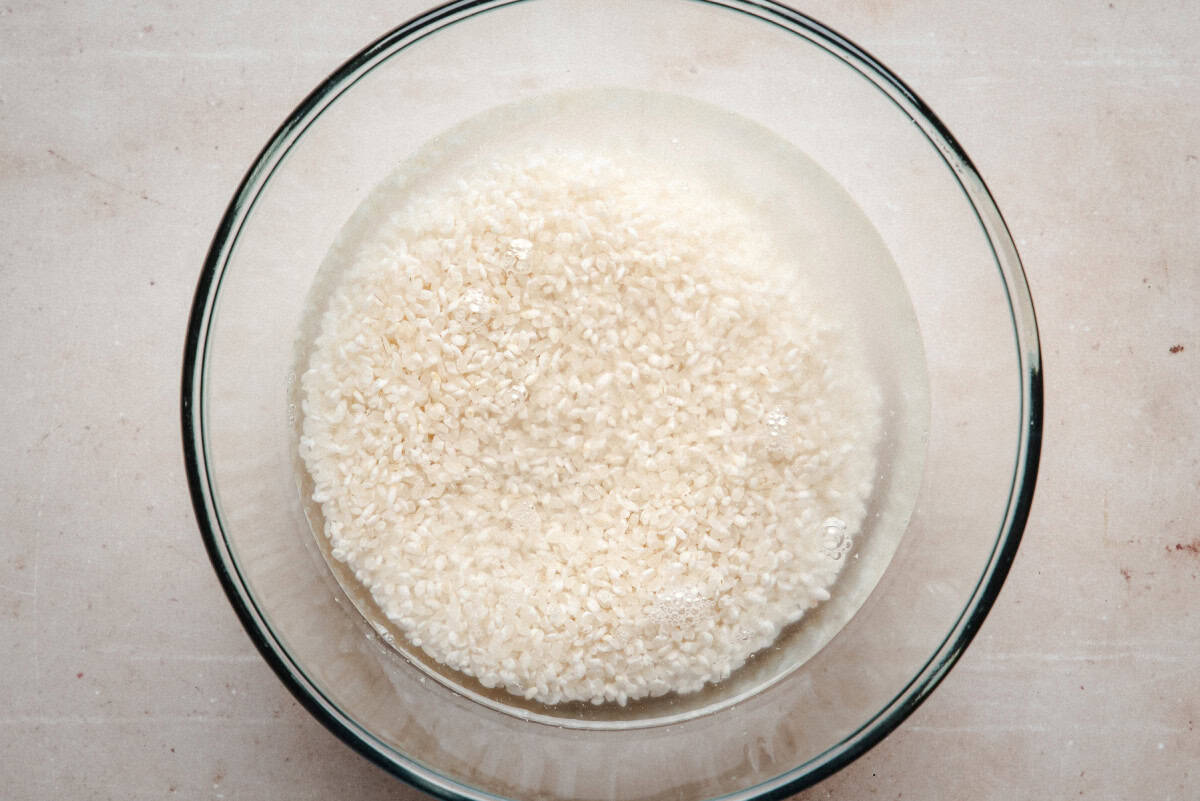 A glass bowl filled with uncooked white rice soaking in water, placed on a light-colored surface.