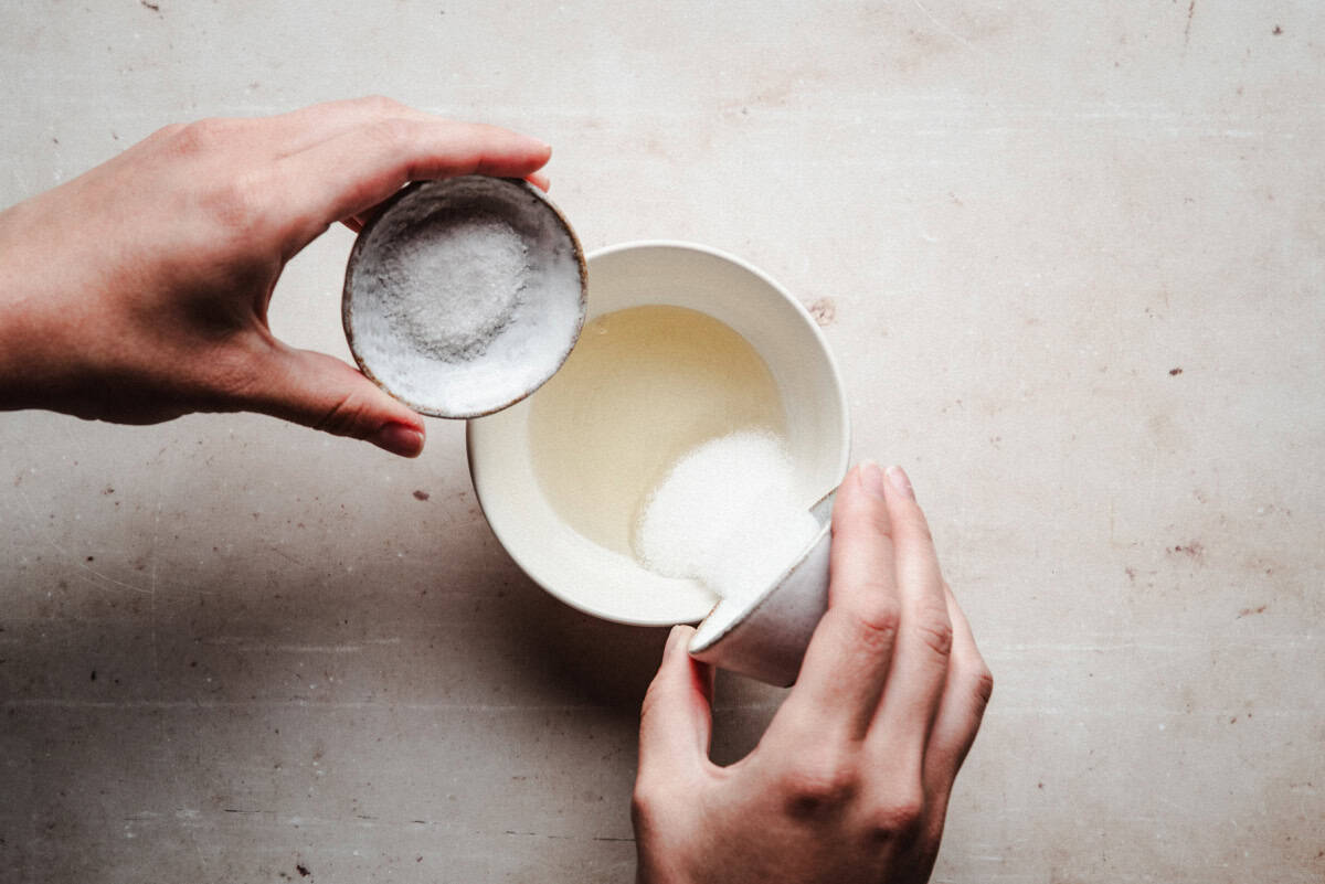 Two hands hold small bowls of salt and sugar, pouring them into a white bowl containing a rice vinegar on a light countertop.
