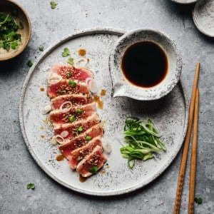 A plate of seared tuna slices topped with green onions and sesame seeds, served with a side of soy sauce, sliced scallions, and wooden chopsticks on a textured gray surface.