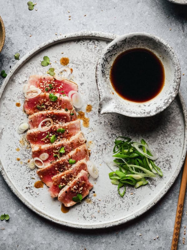 A plate of seared tuna slices topped with green onions and sesame seeds, served with a side of soy sauce, sliced scallions, and wooden chopsticks on a textured gray surface.