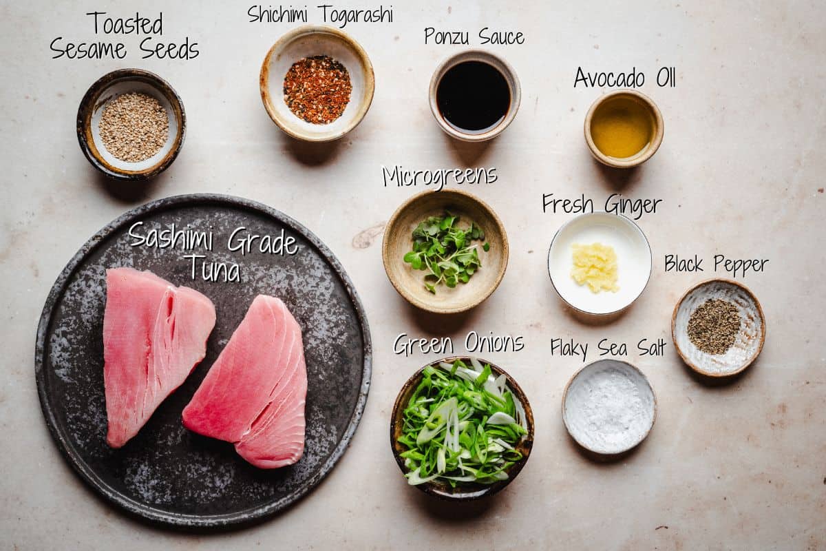 A flat lay of ingredients for tuna tataki, including tuna slices on a plate, toasted sesame seeds, shichimi togarashi, ponzu sauce, avocado oil, microgreens, fresh ginger, green onions, black pepper, and flaky sea salt.