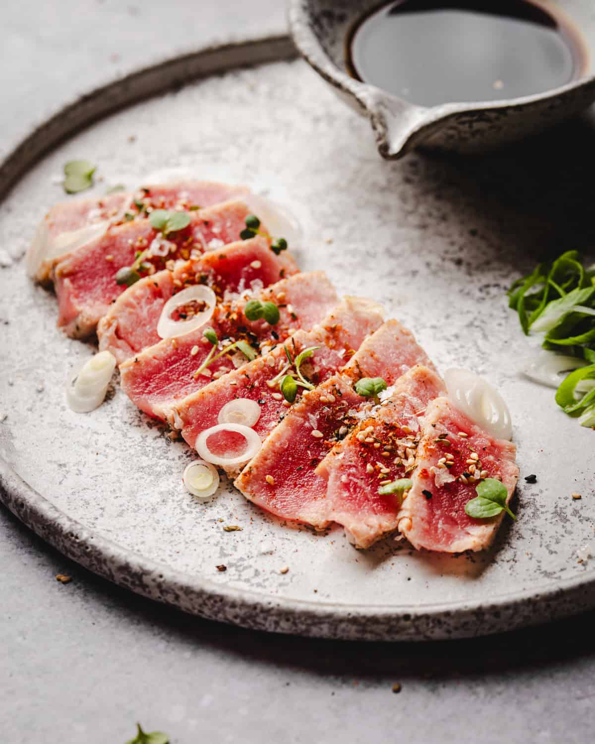 Slices of seared tuna topped with microgreens, thinly sliced onions, and spices are arranged on a round ceramic plate. A small bowl of soy sauce and sliced green onions sit beside the dish.
