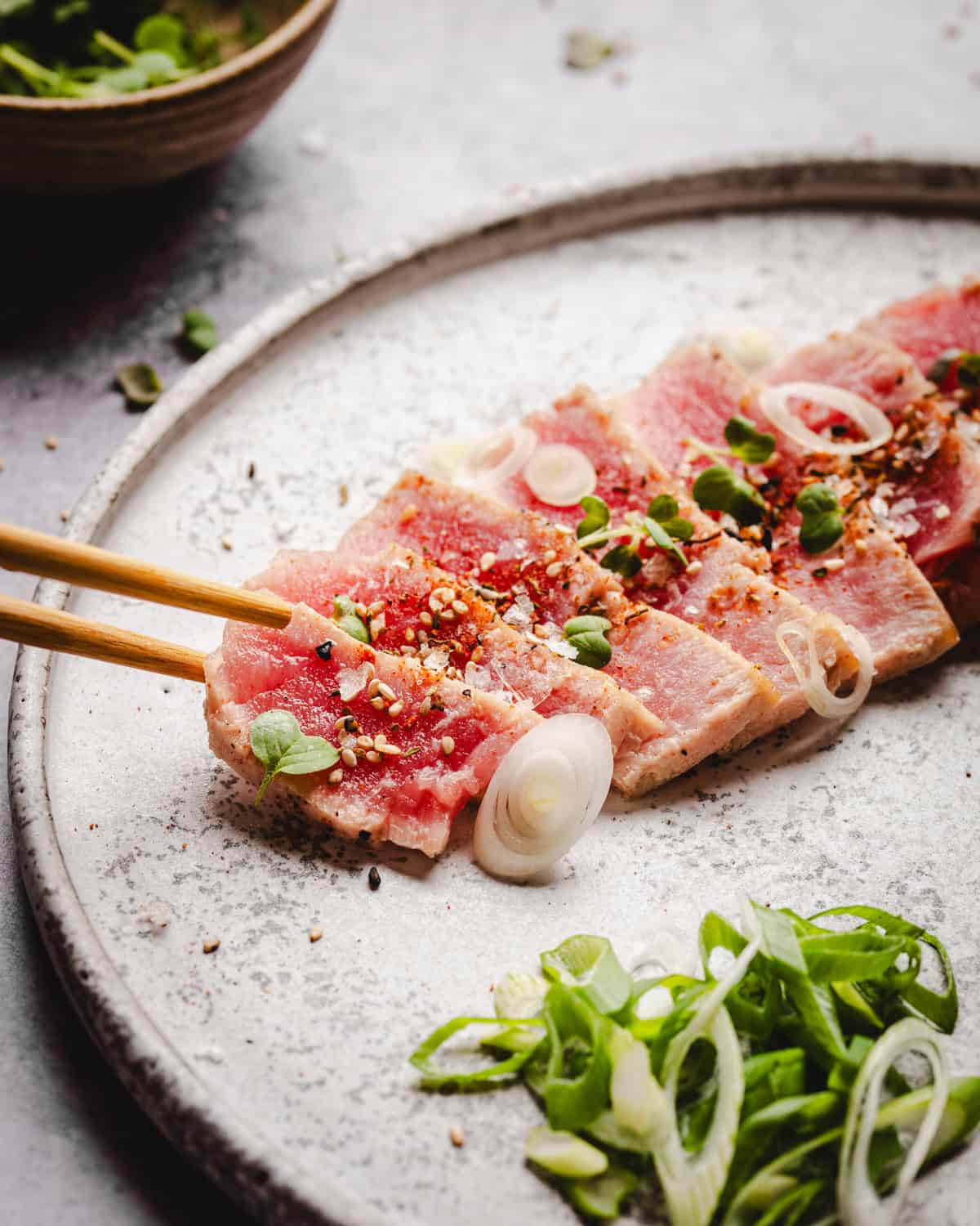 Chopsticks hold a slice of seared tuna tataki topped with sesame seeds, green onions, and spices on a speckled plate. Sliced green onions and microgreens are arranged nearby as garnish.