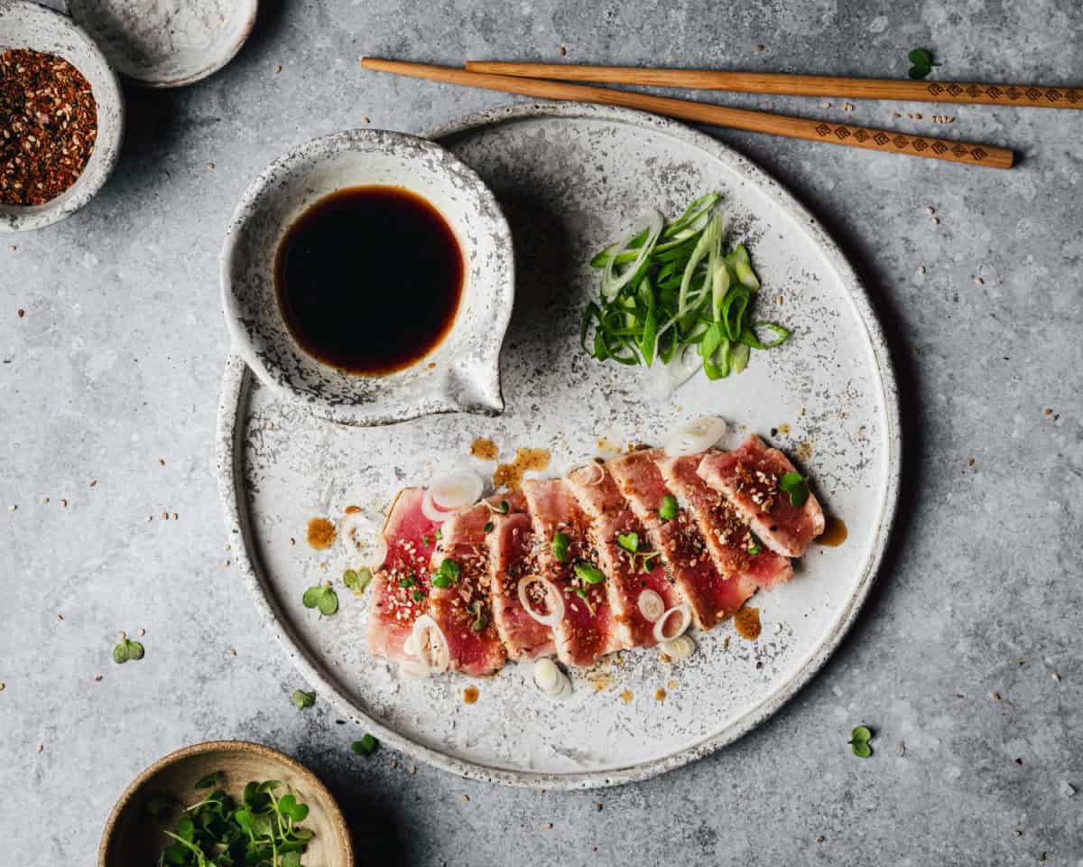 A plate of seared tuna slices topped with green onions and sesame seeds, served with a side of soy sauce and garnished with sliced scallions; chopsticks and small bowls of spices and herbs are nearby.