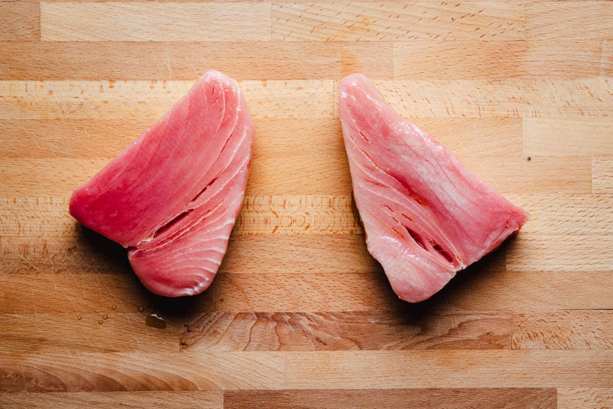 Two raw tuna steaks with a pink hue are placed side by side on a light wooden cutting board, showcasing the texture and marbling of the fish.