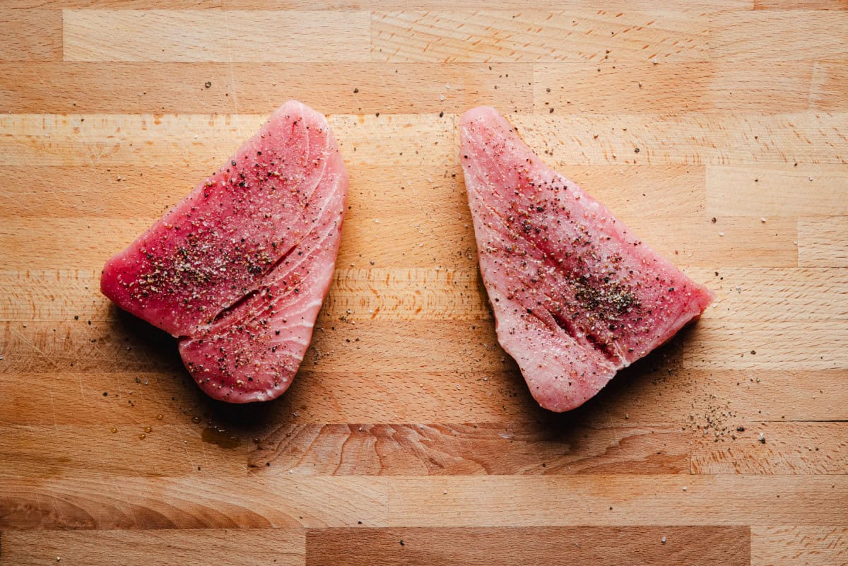 Two raw tuna steaks seasoned with pepper and salt are placed on a wooden cutting board. The pieces are arranged with space between them, showing the texture of both the fish and the board.