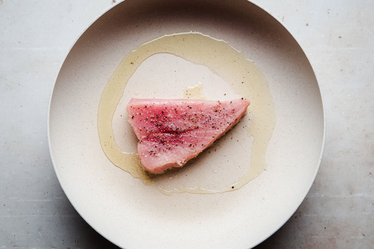 A raw tuna steak sits on a beige pan, seasoned with cracked black pepper and surrounded by a drizzle of olive oil.