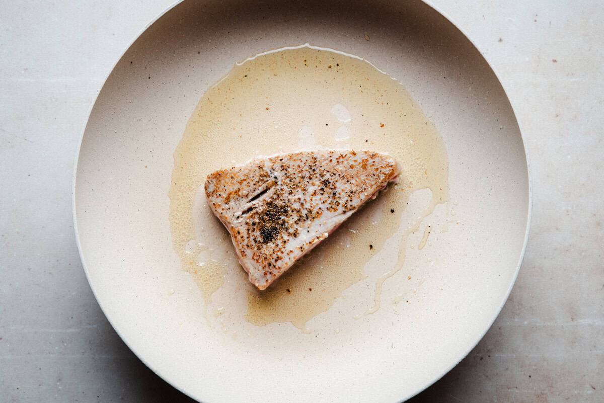 A piece of seared tuna steak seasoned with pepper sits on a beige pan in a small pool of cooking juices. The background and plate are neutral in color, highlighting the simplicity of the dish.