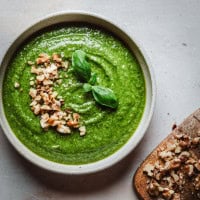 A bowl of walnut pesto garnished with chopped nuts and fresh basil leaves, next to a wooden board with more chopped nuts on a light surface.