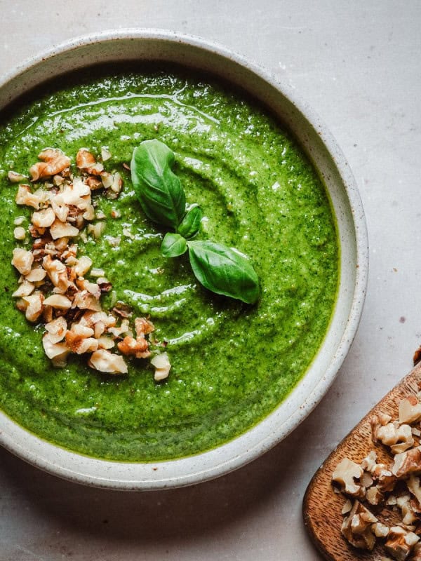 A bowl of walnut pesto garnished with chopped nuts and fresh basil leaves, next to a wooden board with more chopped nuts on a light surface.