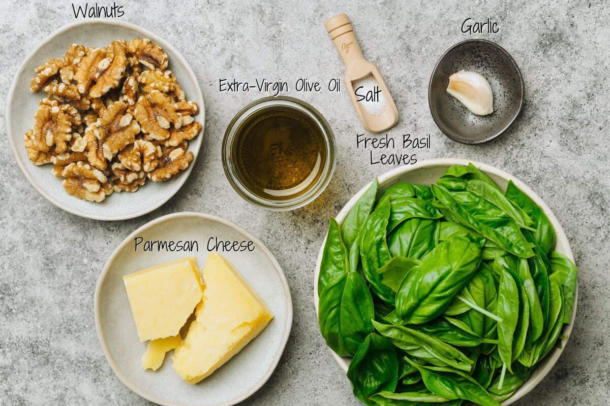 Five bowls on a gray surface containing walnuts, a jar of extra-virgin olive oil, salt with a garlic clove, Parmesan cheese, and fresh basil leaves, each labeled with handwritten text.