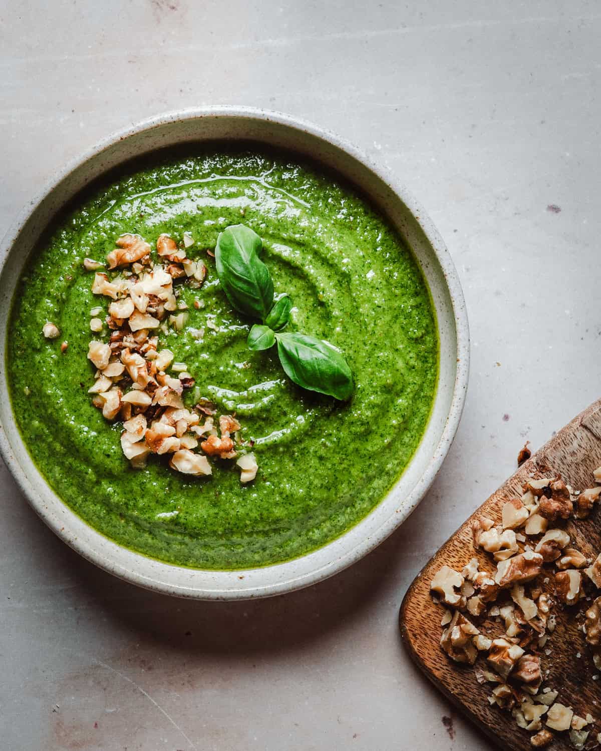 A bowl of walnut pesto topped with chopped nuts and two basil leaves, beside a wooden board with more chopped nuts, on a light-colored surface.