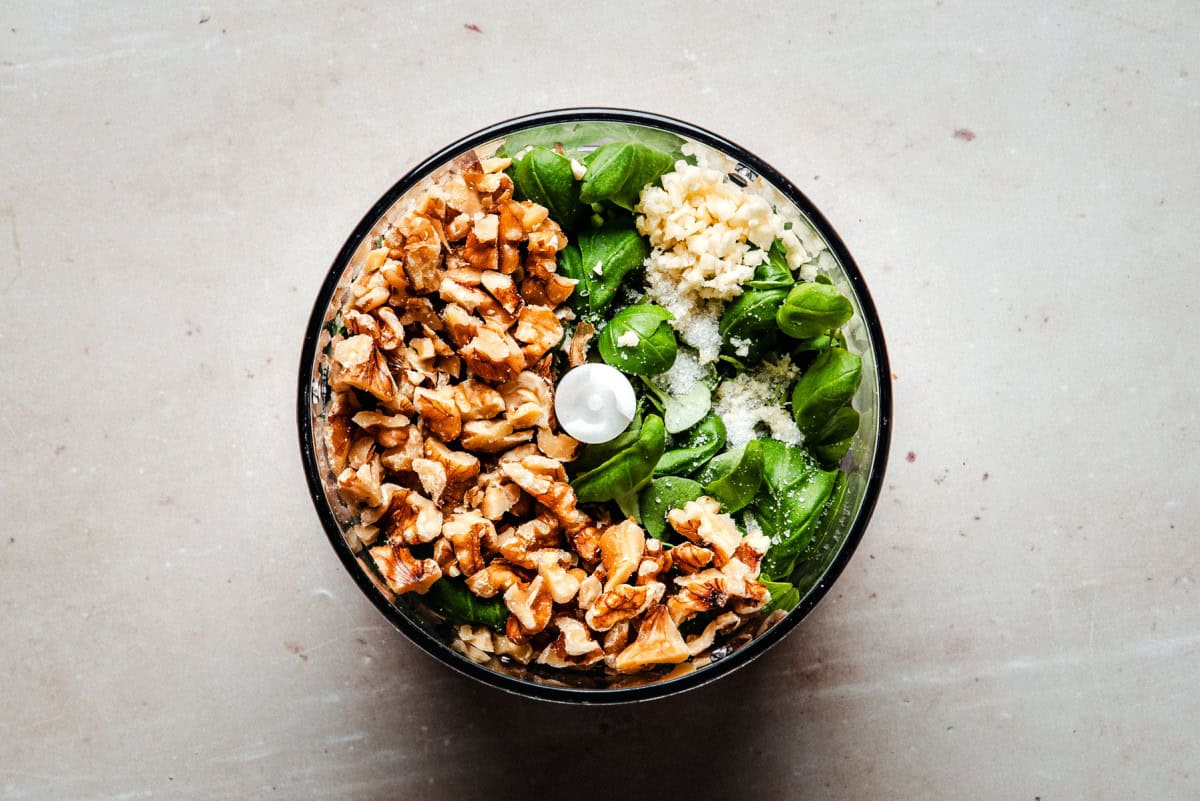 A food processor bowl filled with fresh basil leaves, chopped walnuts, grated cheese, and minced garlic, ready to be blended into pesto, photographed from above on a light surface.