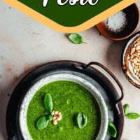 Overhead view of a bowl of green basil pesto garnished with pine nuts and basil leaves, placed on a black tray with scattered basil leaves and a small bowl of pine nuts nearby. Text reads "Fresh Basil Pesto.