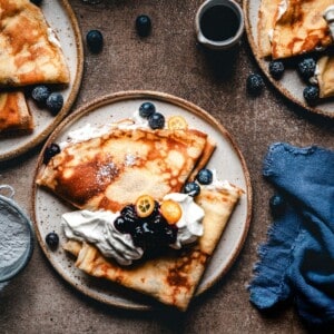 A plate of crepes topped with whipped cream, blueberries, sliced kumquats, and a dark berry sauce, surrounded by additional plates of crepes, a cup of syrup, and a blue cloth napkin.