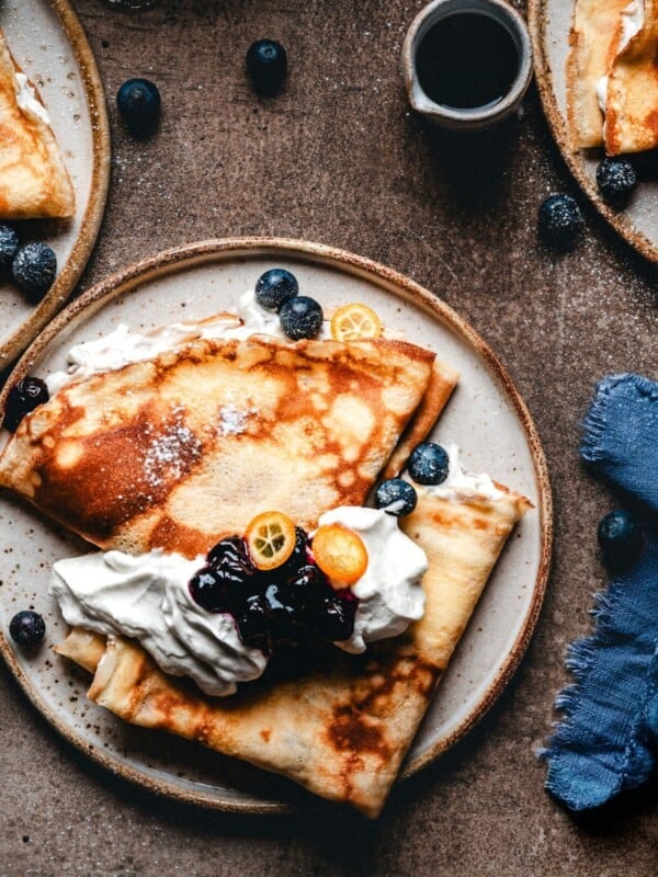 A plate of crepes topped with whipped cream, blueberries, sliced kumquats, and a dark berry sauce, surrounded by additional plates of crepes, a cup of syrup, and a blue cloth napkin.